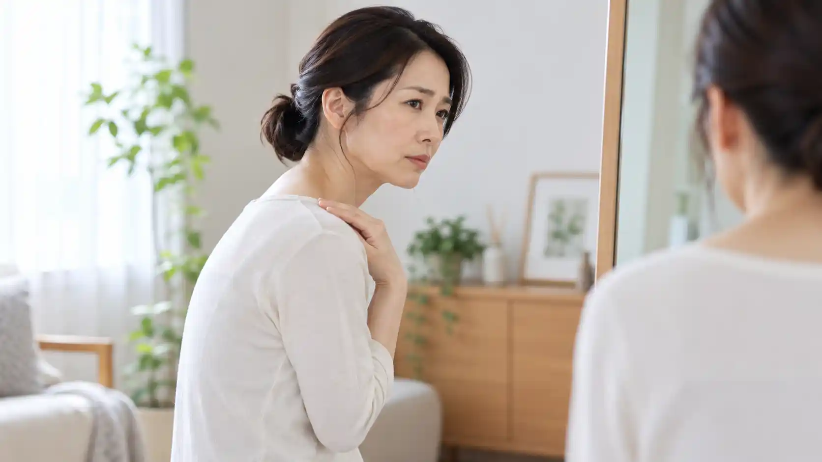 鏡の前で首を少し前に傾け、手を肩に添えながら首や肩の状態を確認している女性。