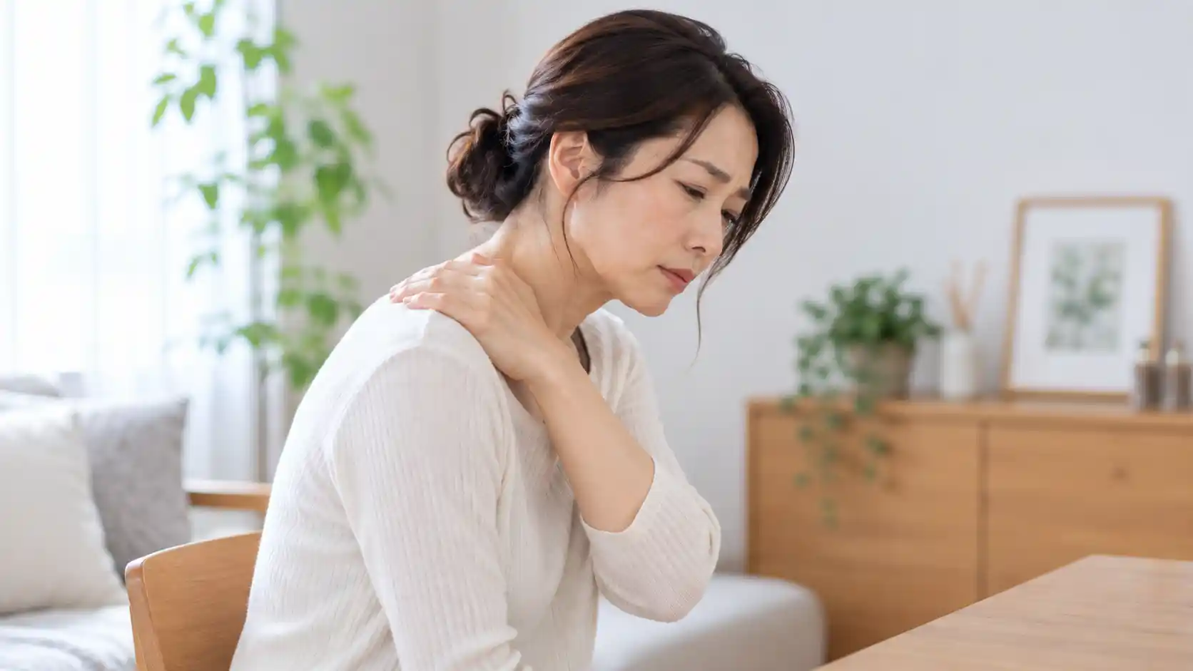 椅子に座り、首を前に傾けながら片手を肩に添えて、首や肩の痛みやこりを気にしている女性。