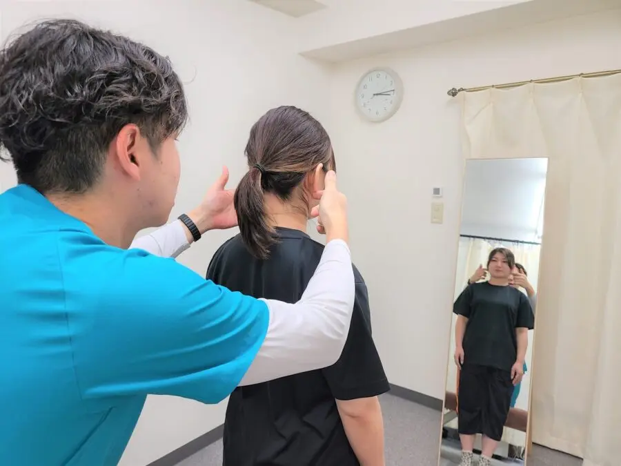 自由が丘あゆむ整体院で、院長が鏡の前に立つ女性のお客様の頭や首の位置を指し示しながら、姿勢や身体のバランスを確認している様子。