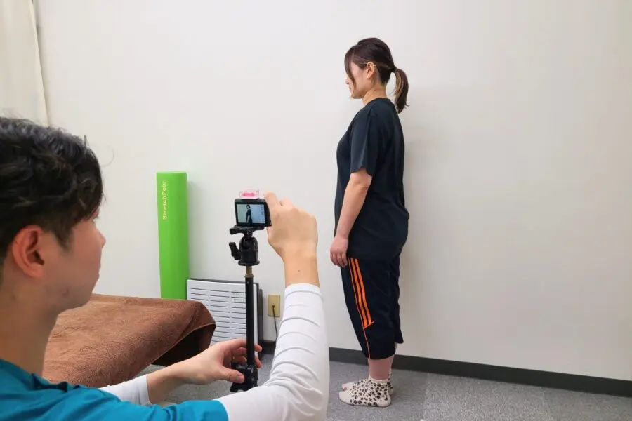 女性が壁の前に横向きで立ち、施術者が三脚に設置したカメラを使用して、身体の姿勢を横から撮影している様子。