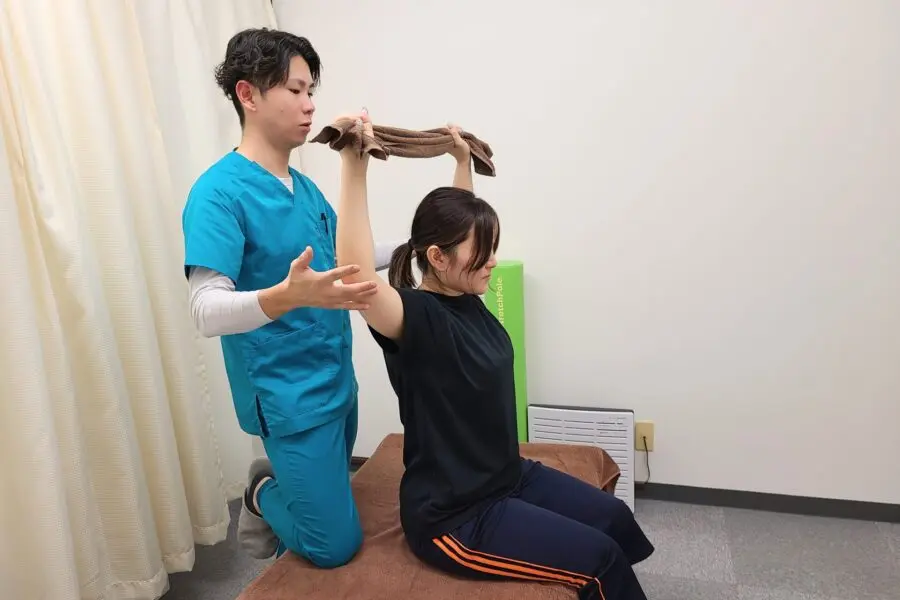 施術ベッドに座った女性がタオルを両手で持ち、腕を頭上に上げて肩関節の動きを確認している様子。施術者が隣で動作を観察している。