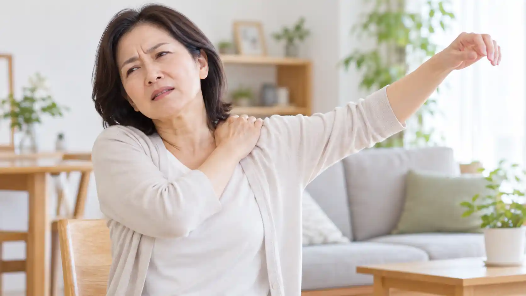 椅子に座り、片腕を上げながら反対の手で肩を押さえ、痛そうな表情を浮かべている女性。