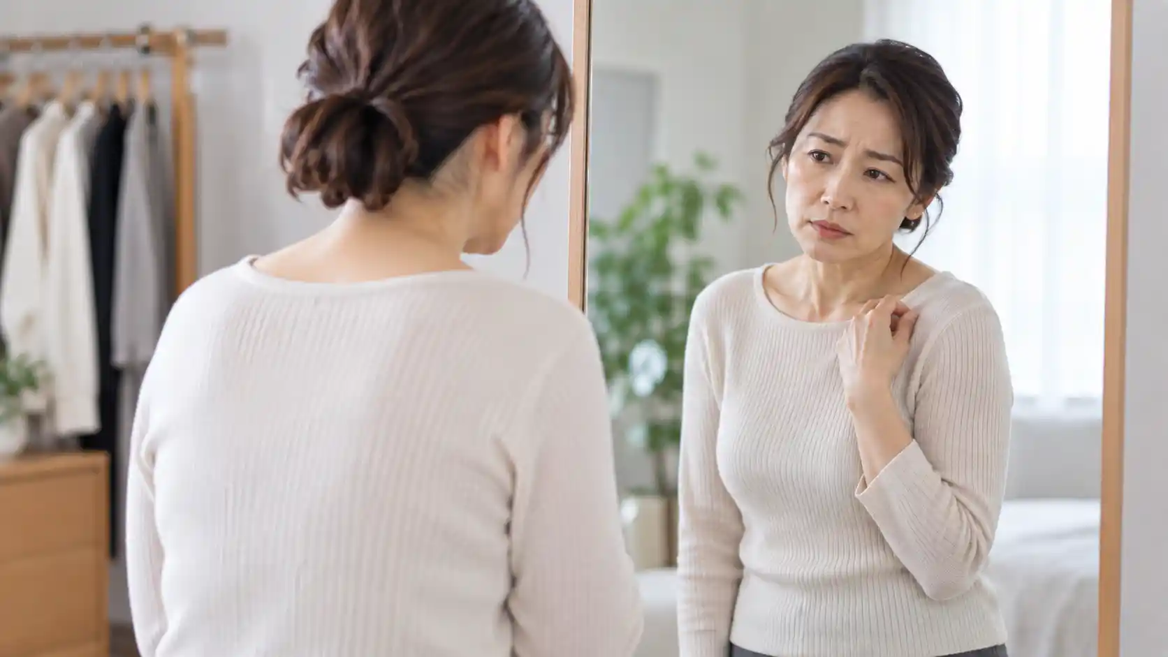 鏡の前に立ち、片手を肩に添えながら、自分の肩の位置や姿勢を気にしている女性。