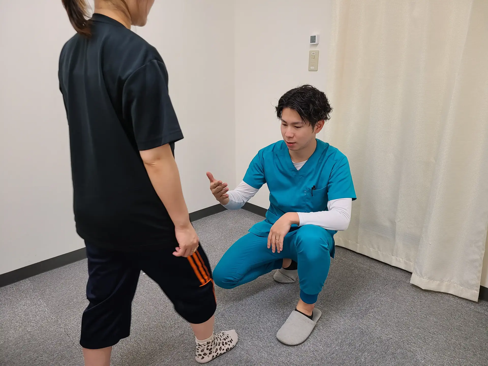 自由が丘あゆむ整体院で、青い施術着を着た院長が片膝をつき、立っている女性のお客様の脚の位置や立ち方を確認している様子。
