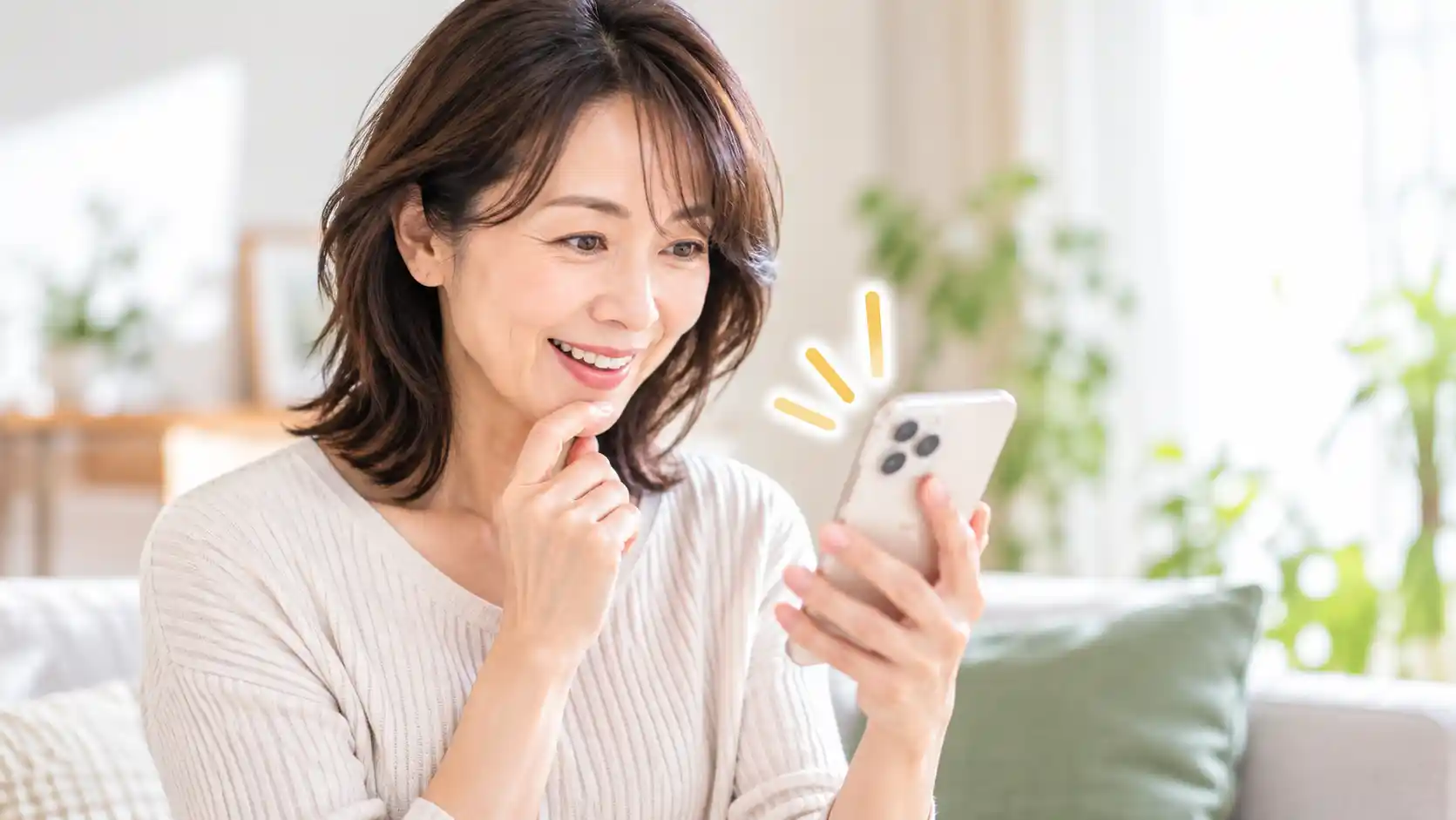 明るい室内でソファに座り、スマートフォンの画面を見ながら笑顔を浮かべている女性。